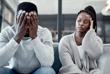 Woman sitting on a couch looking frustrated while a man covers his face with his hands after an argument.
