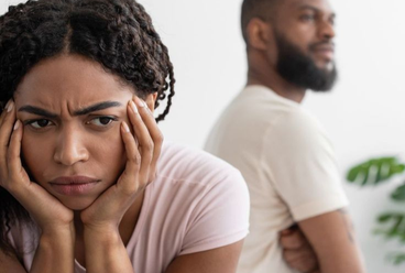Woman sitting with her head in her hands looking upset while a man stands in the background turned away after an argument.