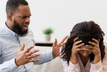 Man angrily yelling while a woman sits with her head in her hands, visibly overwhelmed and distressed.