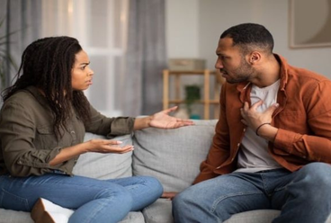 Couple sitting on a couch having an intense argument, with the woman gesturing in frustration and the man defending himself.
