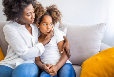 Woman sitting on a couch with a young girl, her hand gently resting on the child’s face.