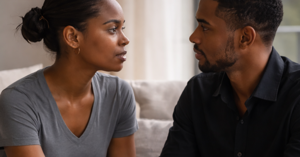 A couple sits closely on a sofa, holding hands and making steady eye contact while having a serious conversation in a softly lit living room.